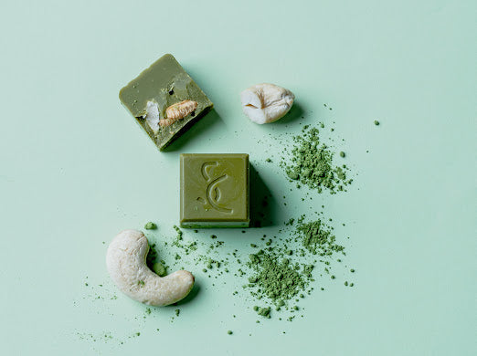 Cube of cacao chocolate carrying Sacred Cacao emblem surrounded by green powder and cashew nuts.